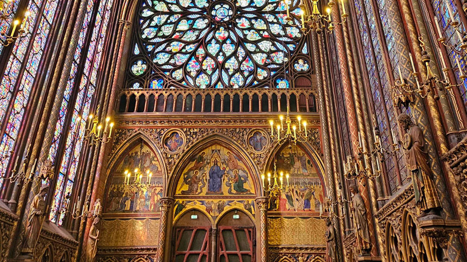 Vue intérieure du fond de la chapelle haute de la Sainte-Chapelle à Paris, avec une grande rosace gothique au-dessus d’arcades richement décorées, de scènes peintes, de sculptures dorées et de hautes verrières.